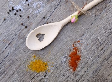 Cuillère de bois percée d'un coeur au centre et une mini rose attaché au manche, avec des épices, le tout sur un comptoir de bois.
